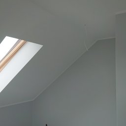 Robert Stachowiak StachBUD - Poddasze z oknem dachowym po świeżym pomalowaniu na szaro, widoczna listwa przypodłogowa i wystający kabel ze ściany.