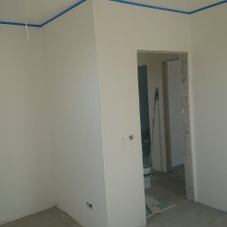 Robert Stachowiak StachBUD - Przygotowania do malowania w nowym domu: ściany zagruntowane i zabezpieczone niebieską taśmą malarską przy suficie, widoczne puszki z farbą oraz narzędzia w przejściu do innego pomieszczenia...