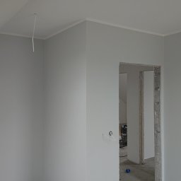 Robert Stachowiak StachBUD - Świeżo pomalowane na szaro wnętrze poddasza z oknem dachowym, widoczne ślady po gładzi na ścianach i sufitach, oraz surowy otwór drzwiowy z widocznym murem.