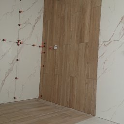 Robert Stachowiak StachBUD - Układanie płytek imitujących drewno na ścianie i podłodze w łazience, z użyciem systemu poziomowania płytek (klipsy i kliny), widoczne przewody elektryczne wystające z sufitu.