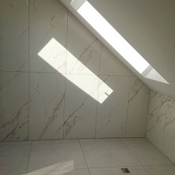 Robert Stachowiak StachBUD - Wnętrze łazienki na poddaszu z oknem dachowym, wykończone płytkami imitującymi marmur w odcieniach bieli i beżu, z widocznymi elementami instalacji wodnej i fragmentem niedokończonej ściany.