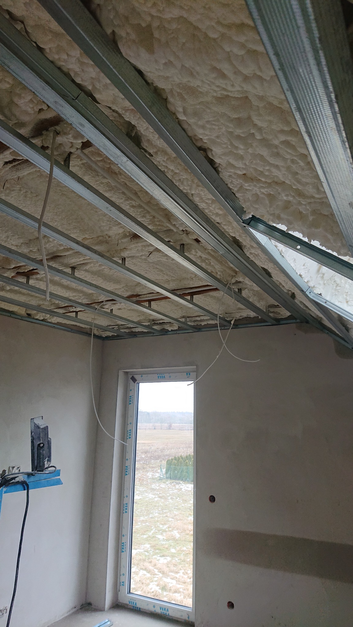 Poddasze w trakcie izolacji, widoczne metalowe profile konstrukcyjne sufitu podwieszanego z wypełnieniem z pianki izolacyjnej, okno z widokiem na zimowy krajobraz, ściany w stanie surowym.