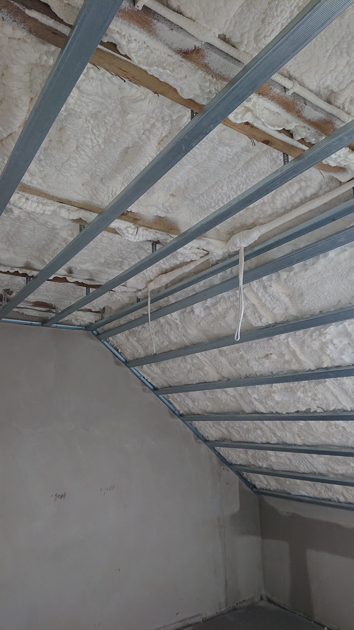 Poddasze w trakcie izolacji pianą, widoczne metalowe profile konstrukcji pod płyty gipsowo-kartonowe, surowe ściany w tle.