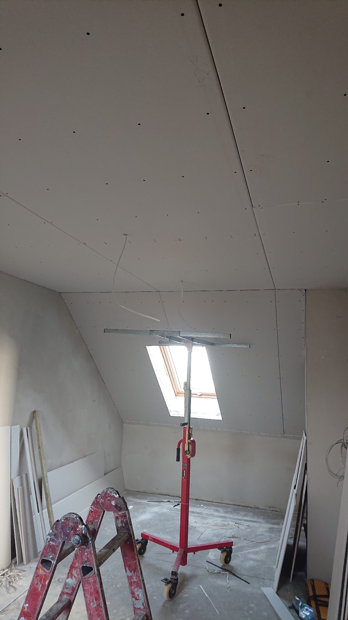 Poddasze w trakcie prac wykończeniowych z zamontowanymi płytami gipsowo-kartonowymi na suficie i ścianach, widoczne okno dachowe, podnośnik do płyt GK i drabina malarska.