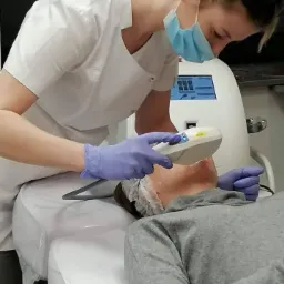 Zabieg na twarz pacjentki wykonywany przez kosmetologa w białym uniformie i niebieskich rękawiczkach, z użyciem specjalistycznego urządzenia.