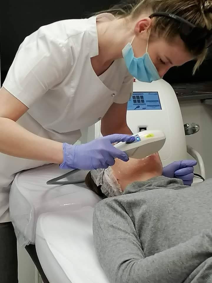 Zabieg na twarz pacjentki wykonywany przez kosmetologa w białym uniformie i niebieskich rękawiczkach, z użyciem specjalistycznego urządzenia.