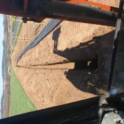 Wąski wykop w ziemi z ułożonymi czarnymi rurami, widok z perspektywy maszyny budowlanej na tle pola i drzew.