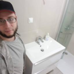 Paweł Andronczyk - Mężczyzna w okularach i czapce robi selfie w łazience po remoncie, widoczna umywalka z szafką, bateria, dozownik mydła oraz kabina prysznicowa z odpływem liniowym.