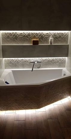 Nowoczesna łazienka z narożną wanną obudowaną mozaiką, podświetlane półki z dekoracyjnymi płytkami i subtelnym oświetleniem LED pod wanną i półkami.