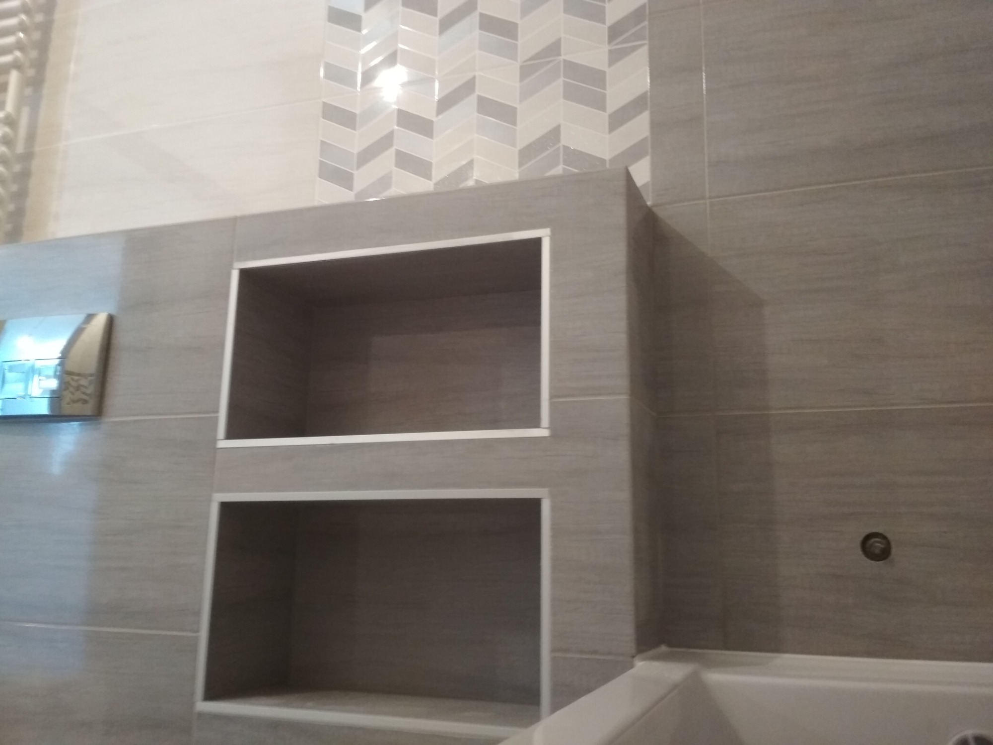 Szare płytki w łazience z wnękami wykończonymi srebrną listwą, dekoracyjny pas płytek w jodełkę, widoczny fragment wanny