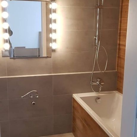 Nowoczesna łazienka z szarymi płytkami, podświetlanym lustrem i wanną obudowaną drewnem.