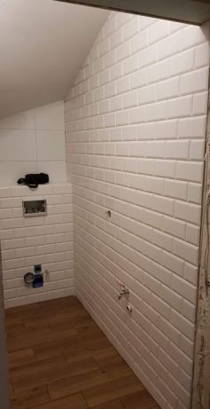 Łazienka w trakcie remontu: białe, wypukłe płytki ułożone na ścianach, widoczne instalacje wodne i elektryczne, drewnopodobna podłoga z paneli, sufit ze skosem.