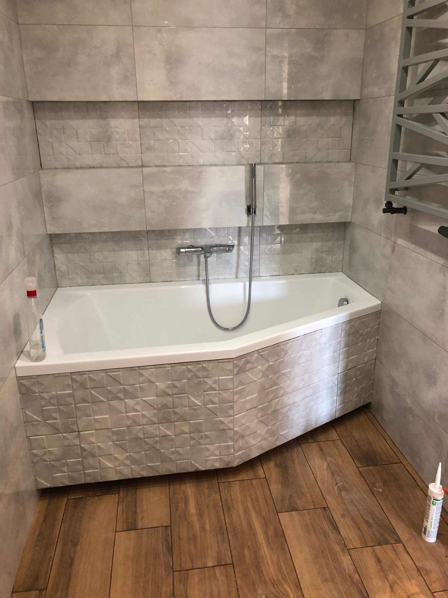 Biała wanna narożna obłożona szarymi płytkami 3D, szare płytki ścienne w dużym formacie, drewnopodobna podłoga, grzejnik drabinkowy i butelka płynu do czyszczenia w tle.