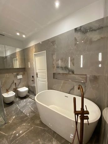 Elegancka łazienka z marmurową posadzką i ścianami, wyposażona w wolnostojącą białą wannę, bidet i toaletę, z akcentami w kolorze miedzianym.