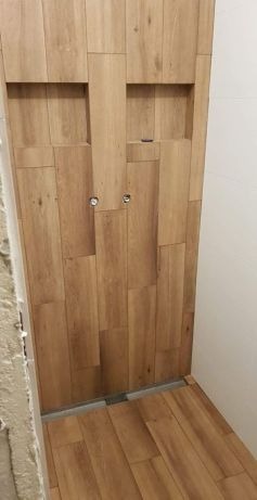 Wykończona ściana prysznicowa z drewnopodobnych płytek ceramicznych ułożonych w nieregularny wzór z wnękami na kosmetyki i widocznymi przyłączami wodnymi.