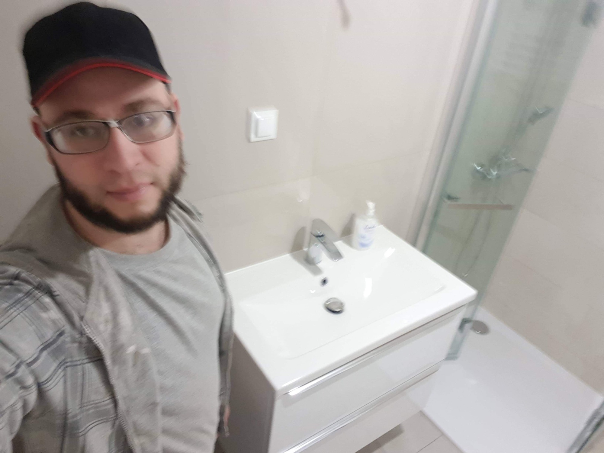 Mężczyzna w okularach i czapce robi selfie w łazience po remoncie, widoczna umywalka z szafką, bateria, dozownik mydła oraz kabina prysznicowa z odpływem liniowym.