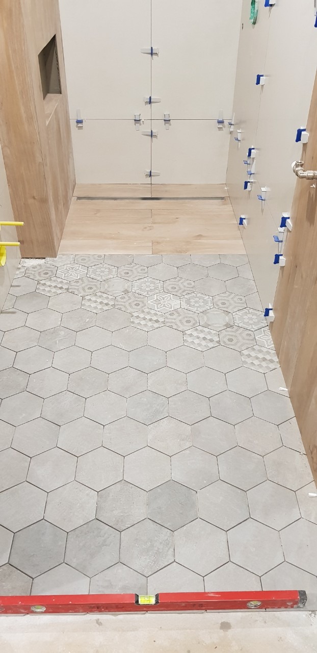 Układanie szarych, heksagonalnych płytek podłogowych w łazience, częściowo z dekoracyjnym wzorem, z poziomnicą czerwoną w dolnej części kadru i widocznymi krzyżykami dystansowymi na ścianie.