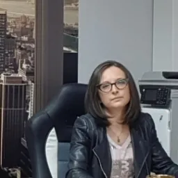 Portret kobiety w okularach siedzącej na krześle biurowym, w tle widok na miasto i urządzenie biurowe.