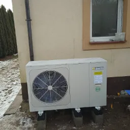 Zewnętrzna jednostka pompy ciepła Beretta zamontowana na betonowych podstawach przy elewacji domu, widoczne instalacje i otoczenie z resztkami śniegu.