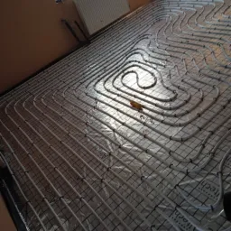 Rozłożone rury ogrzewania podłogowego na folii izolacyjnej z rastrem, widoczne mocowania i narzędzie montażowe w trakcie instalacji.