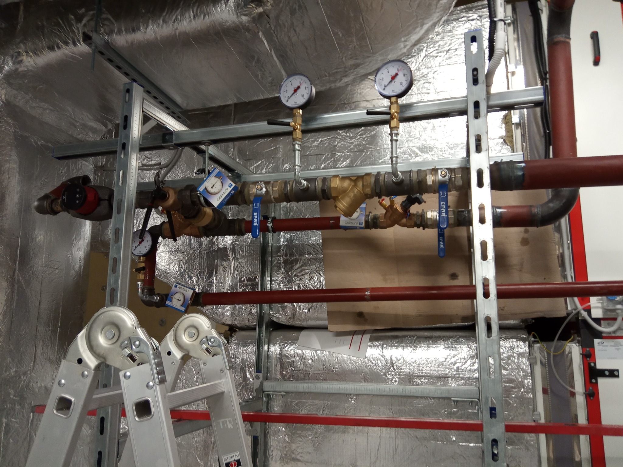 Fragment instalacji grzewczej z manometrami, zaworami i rurami miedzianymi, zamontowanej na konstrukcji wsporczej, widoczna drabina aluminiowa w pomieszczeniu z izolacją.