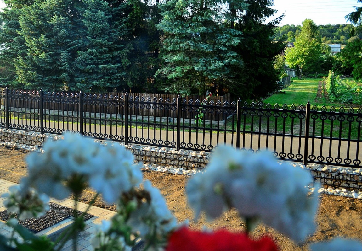 Eleganckie, czarne ogrodzenie metalowe z dekoracyjnymi elementami w kształcie serc, osadzone na kamiennej podmurówce, widoczne zza kwiatów w doniczce, z zielonym ogrodem i drzewami w tle.