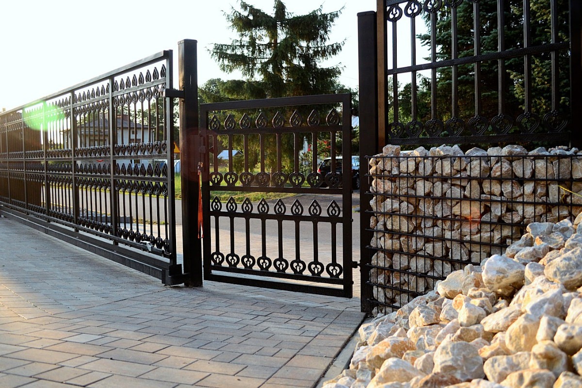 Otwarta, przesuwna, metalowa brama z ozdobnymi, ażurowymi elementami, sąsiadująca z gabionem wypełnionym kamieniami, na tle kostki brukowej i zieleni.