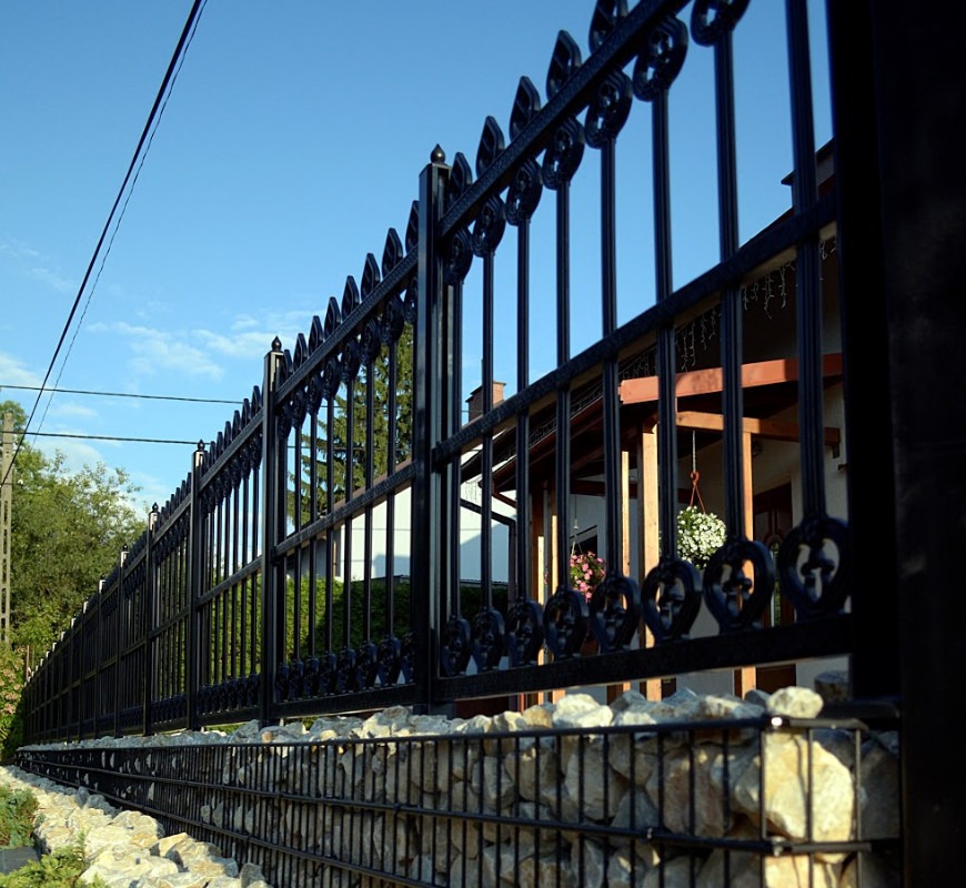 Długie, czarne, metalowe ogrodzenie z dekoracyjnymi elementami w górnej części, osadzone na kamiennym murku gabionowym, na tle domu i błękitnego nieba.
