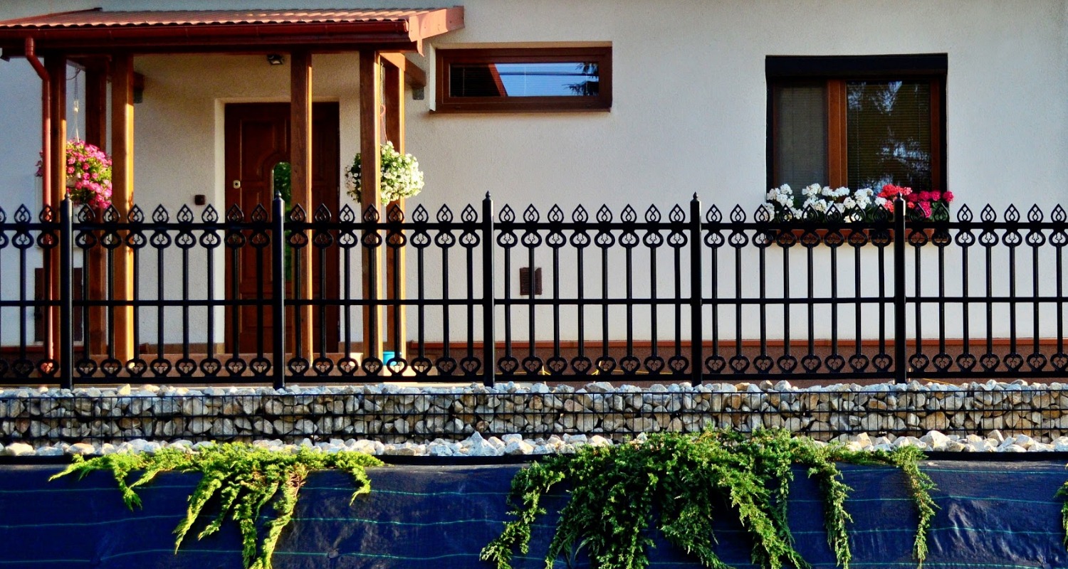 Czarny metalowy płot z ozdobnymi elementami w kształcie serc, oddzielający posesję od skarpy obsadzonej zielenią, z widocznym fragmentem domu z drewnianym gankiem i oknami z kwiatami.