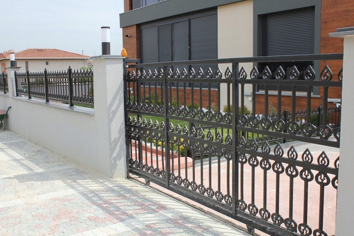 Czarna, metalowa brama wjazdowa i ogrodzenie z dekoracyjnymi elementami w kształcie płomieni, zamontowane na niskim murku z betonowymi słupkami zwieńczonymi lampami, na tle nowoczesnego domu...
