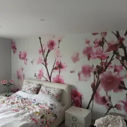 Sypialnia z fototapetą w kwiaty wiśni, łóżko z kwiecistą pościelą, biała szafka nocna i lampa wisząca.