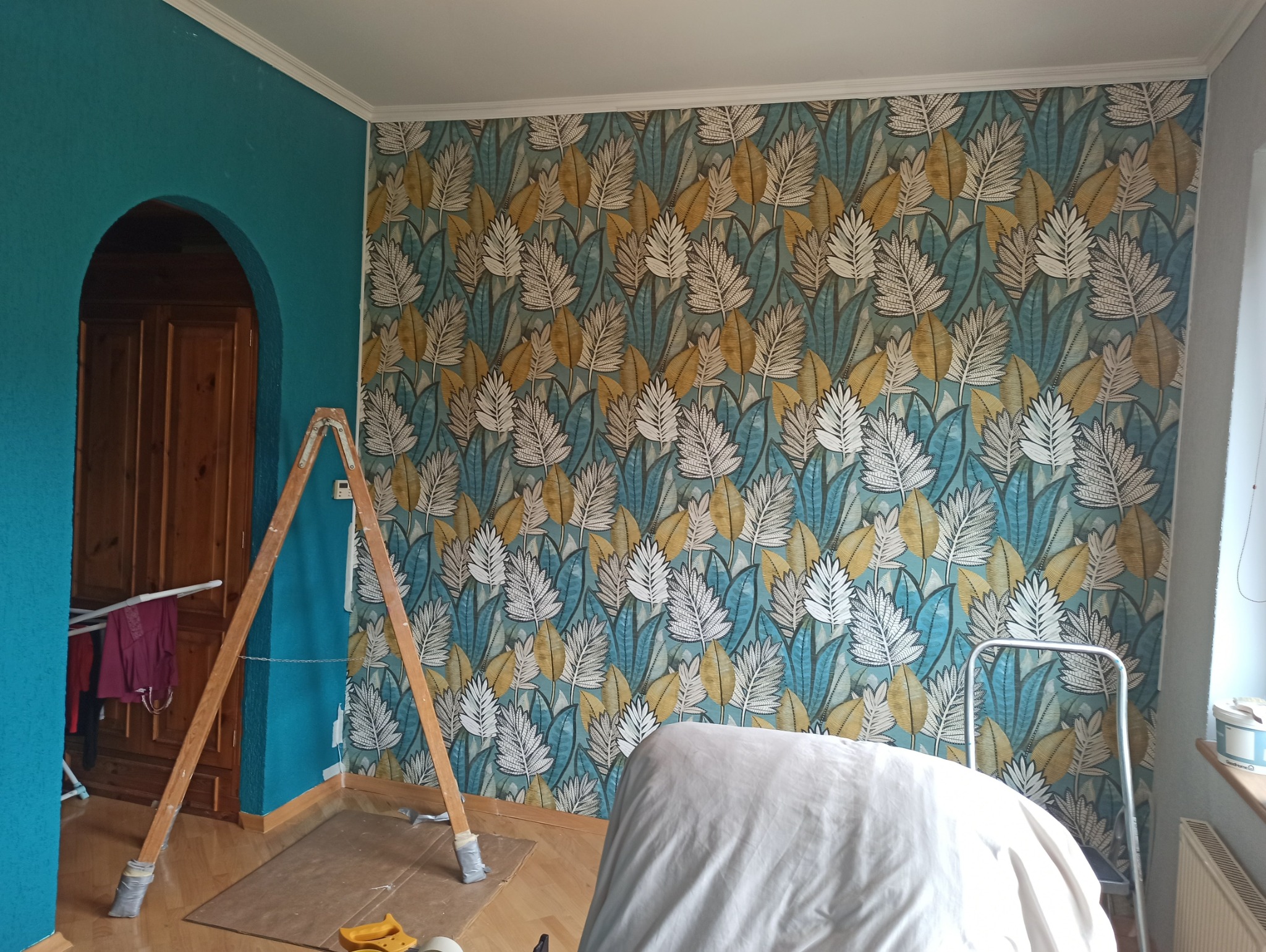 Pokój po tapetowaniu: ściana w liście w odcieniach błękitu, bieli i złota, drabina drewniana oparta o ścianę, fragment turkusowej ściany obok łuku drzwiowego