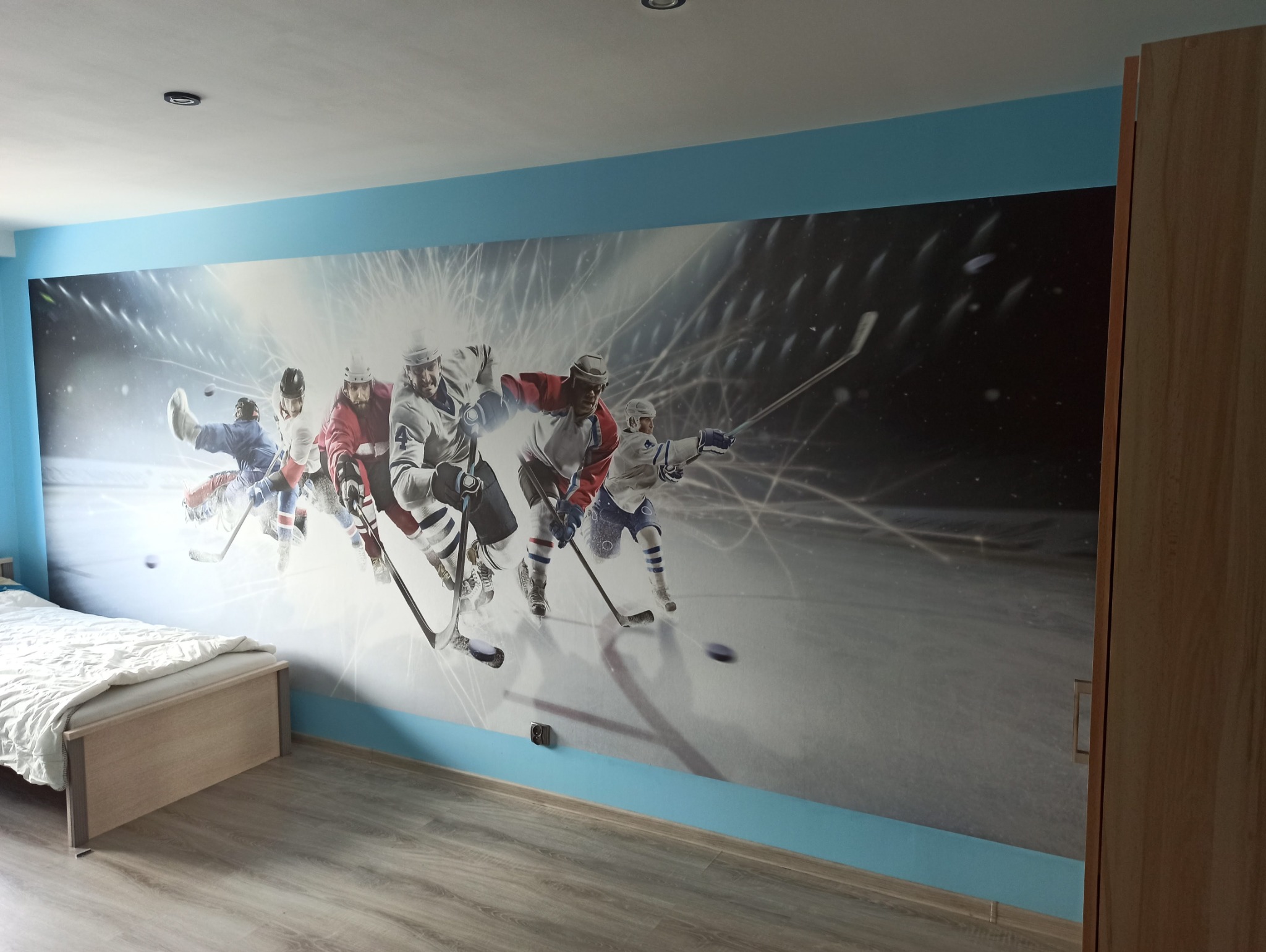 Pokój z fototapetą przedstawiającą dynamiczną scenę hokejową, łóżko z pościelą po lewej stronie, drewniana szafa po prawej.