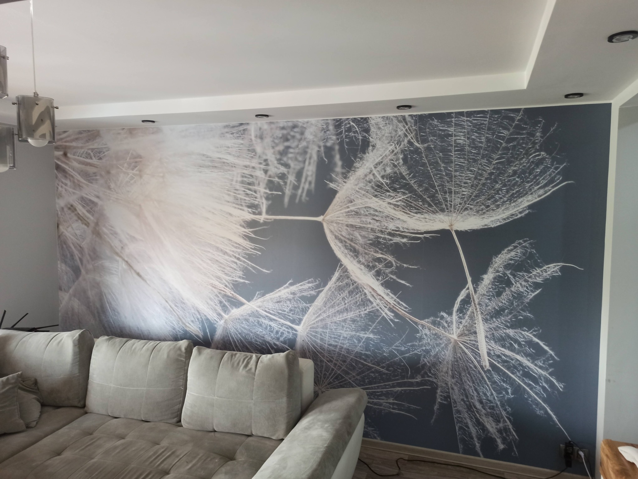 Salon z szarym narożnikiem i fototapetą przedstawiającą dmuchawce na ciemnym tle, oświetlony lampami sufitowymi.