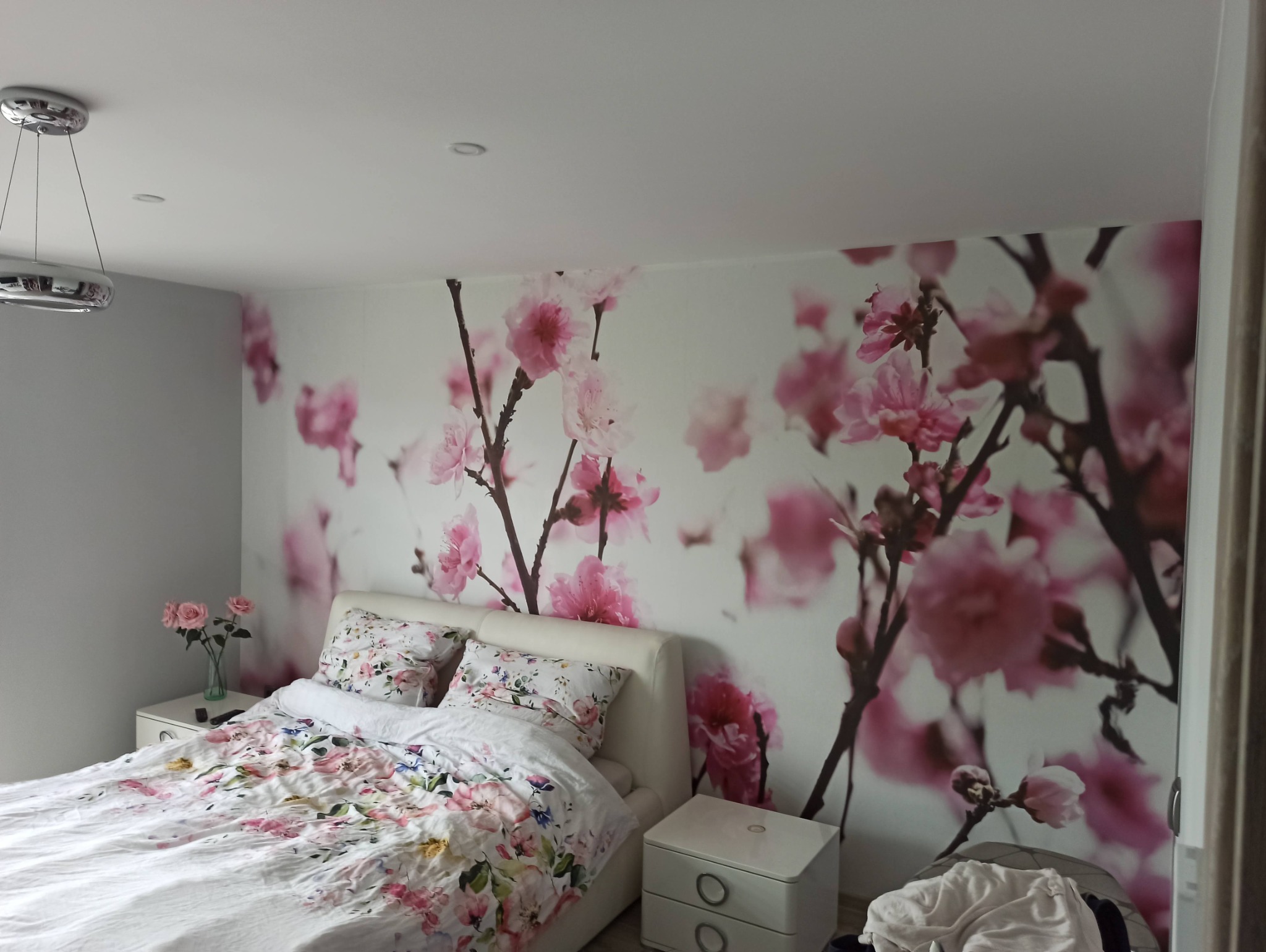 Sypialnia z fototapetą w kwiaty wiśni, łóżko z kwiecistą pościelą, biała szafka nocna i lampa wisząca.