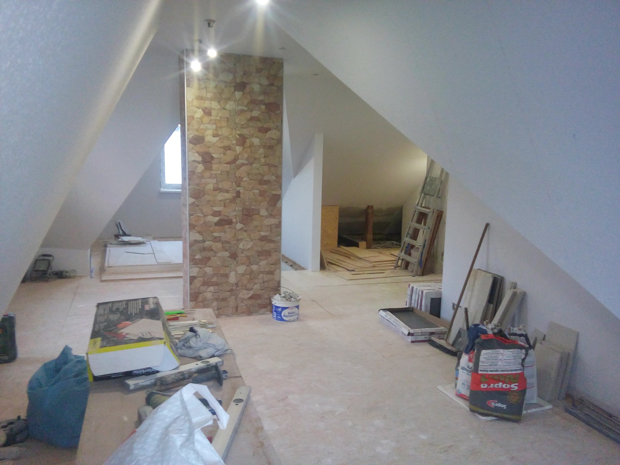Poddasze w trakcie adaptacji: widoczna ściana z dekoracyjnej okładziny kamiennej, materiały budowlane, narzędzia i drabina oparta o ścianę, przygotowane do dalszych prac.