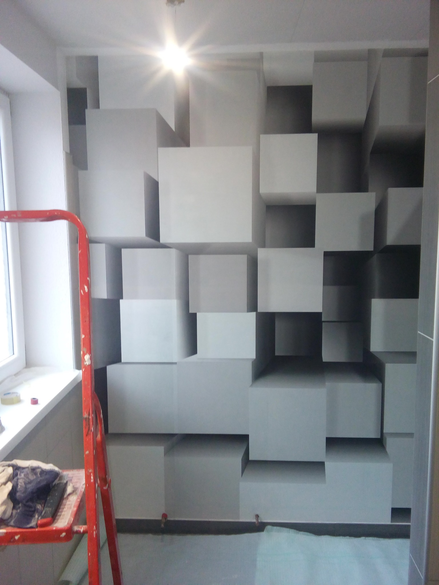 Ściana w pokoju z geometrycznym wzorem 3D z kwadratów w odcieniach szarości, widoczna drabina malarska z resztkami farby i okno z parapetem.