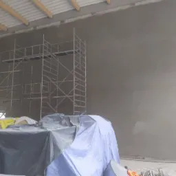 Świeżo otynkowana ściana wewnętrzna z widocznymi rusztowaniami, materiałami budowlanymi przykrytymi folią i odsłoniętą konstrukcją dachu.