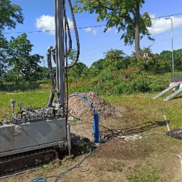 Wiertnica na gąsienicach podczas wiercenia studni głębinowej na zewnątrz, widoczny stos ziemi i narzędzia.