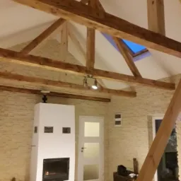 Jasne wnętrze z drewnianą konstrukcją dachu, białym kominkiem z paleniskiem, oknem dachowym i ścianami wyłożonymi dekoracyjną cegłą w odcieniach beżu.