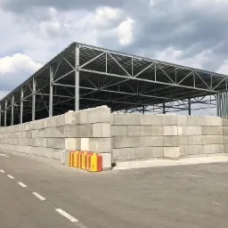 Magazyn prefabrykat&oacute;w betonowych na zewnątrz, ułożonych w wysokie ściany pod metalową konstrukcją dachu, widoczny fragment ciężar&oacute;wki w tle.