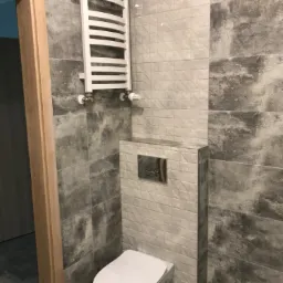 Nowoczesna łazienka z wiszącą toaletą, zabudowaną spłuczką z chromowanym przyciskiem i białym grzejnikiem drabinkowym na ścianie z szarymi płytkami imitującymi kamień.