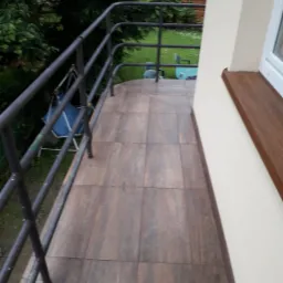 Balkon z brązowymi płytkami imitującymi drewno, szara metalowa balustrada z widokiem na zielony ogród z huśtawką, jasna elewacja budynku z drewnianym parapetem.