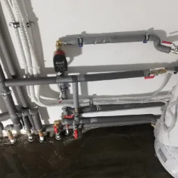 Szare rury instalacji wodnej i grzewczej z zaworami i pompą obiegową Red, zabezpieczone izolacją termiczną, prowadzone wzdłuż białej ściany i ciemnej podłogi. Widoczny fragment owiniętego folią...