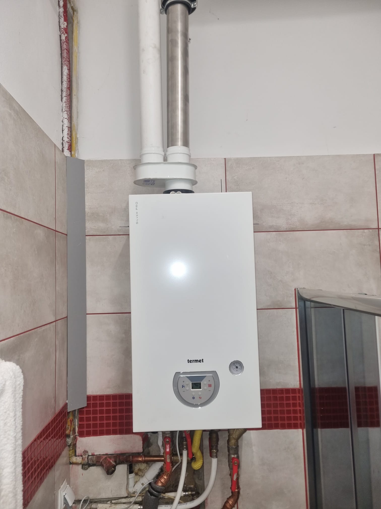 Biały piec gazowy marki Termet, model Silver Pro, zamontowany na ścianie z beżowymi płytkami i czerwoną mozaiką, widoczne rury miedziane i przyłącza, brak estetyki montażu.
