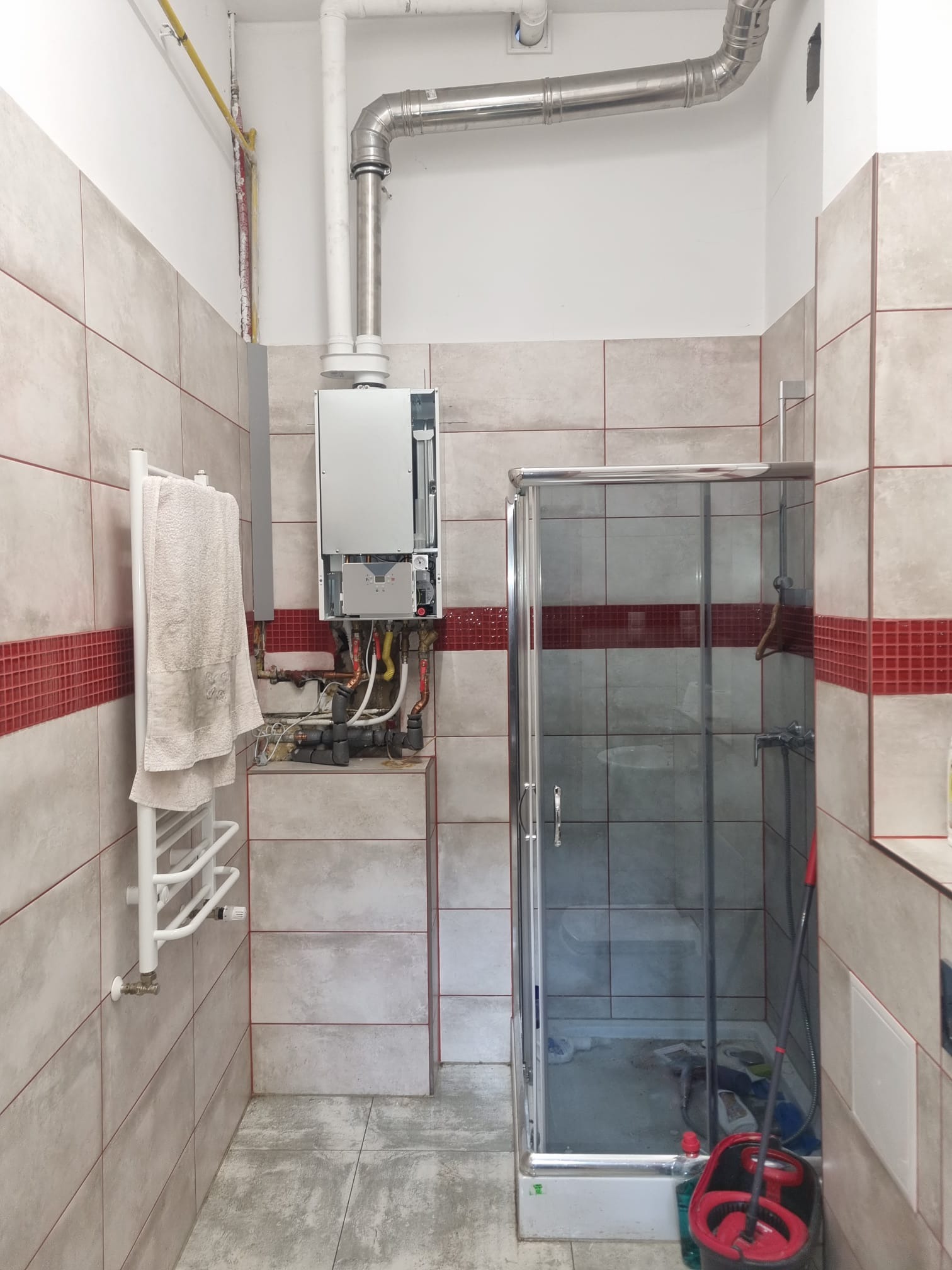 Nowoczesny piec gazowy kondensacyjny zamontowany w łazience z kabiną prysznicową, widoczne rury odprowadzające spaliny i instalacja hydrauliczna.