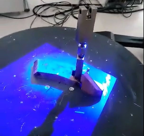 Skan 3D metalowego elementu na obrotowej platformie, oświetlonego niebieskim światłem, z widocznymi przewodami w tle.