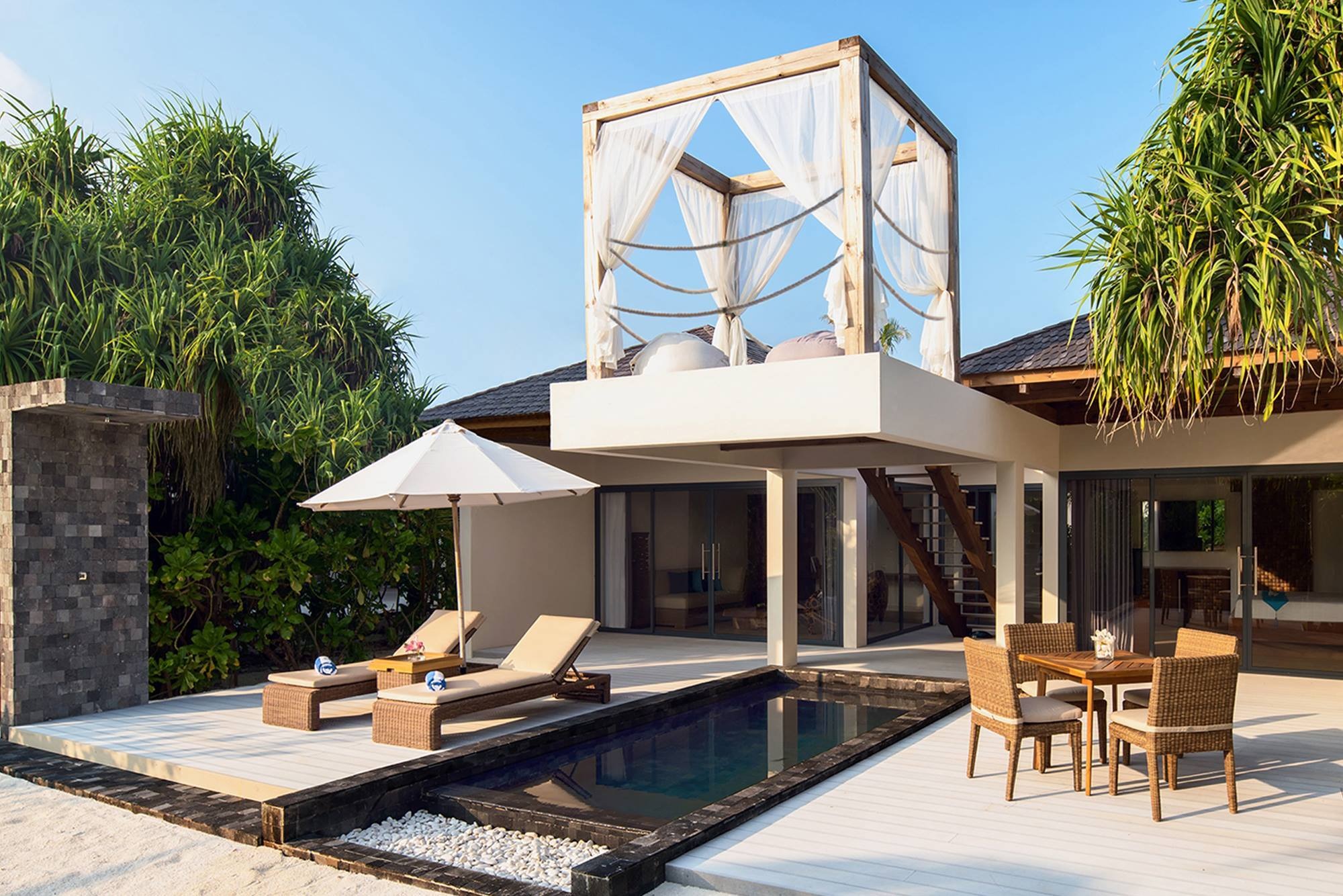 Egzotyczny resort turystyczny z basenem, leżakami pod parasolem i altaną na dachu, otoczony tropikalną roślinnością.