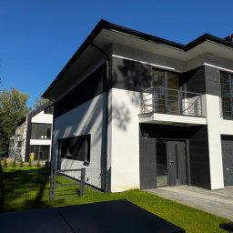 BUDTECHGROUP - Nowoczesny dom z elewacją w odcieniach bieli i grafitu, z widocznymi detalami wykończenia, balkonem i fragmentem ogrodzenia. Na drugim planie dom w trakcie prac elewacyjnych.