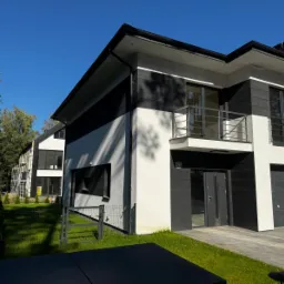 Nowoczesny dom z elewacją w odcieniach bieli i grafitu, z widocznymi detalami wykończenia, balkonem i fragmentem ogrodzenia. Na drugim planie dom w trakcie prac elewacyjnych.